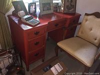 Image showing the full frontal view of the distressed red wooden desk with several drawers and a beige chair next to it.