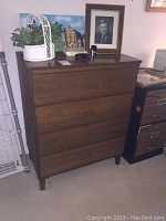 Front angle view of the wooden dresser showing four drawers and decorative metal handles, with some items on top including a framed photo and a plant.