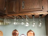 Twenty-nine stemmed glasses hanging inverted from a wooden rack in a kitchen setting, varying in size and shape, including some colored glasses.