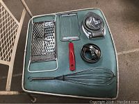 Kitchen utensils including box grater, flat grater with red handle, whisk, egg slicer, biscuit cutters on green chair.