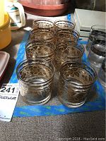 Overall view of the eight gold etched drinking glasses arranged in two rows of four on a table.
