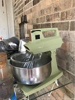 Photo showing vintage Sunbeam stand up mixer in light green with attached large metal mixing bowl on a glass surface against a brick wall.
