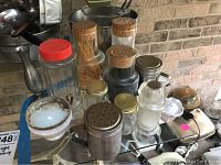 Wide view of assorted vintage glass canisters, jars, and salt and pepper shakers on a clear glass shelf in front of a brick wall.