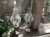 Two glass hurricane candle lamps with decorative etched glass chimneys and metal tripod bases placed side by side on a window ledge.