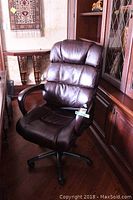 Brown leather office chair with padded segmented backrest and armrests, positioned beside a wooden cabinet and rug on hardwood floor.