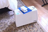 Side view of white carrying case with foot pedal on top, placed on patterned carpet.