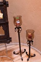 View of both tall metal candle holders with curled feet, decorative rings, and glass hurricane shades holding red candles.