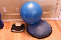 Photo showing an analog weight scale, a large blue exercise ball, and a blue balance trainer (bouncer) on hardwood floor against beige wall.