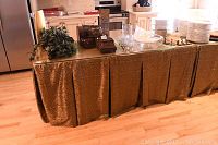 Full view of the folding table covered with a brown and gold patterned fabric skirt, glass top visible to the right side. The table is set in a kitchen or dining area with hardwood floor and cabinetry in background.