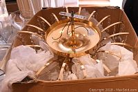 Overhead view of gold-tone metal ceiling fixture in cardboard box showing radial arms and wiring.