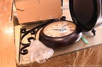 Clock laying flat on table showing the dark bronze round frame, roman numeral display, and ornate decorative metal bracket