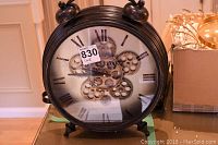 Front view of round black metal gear clock showing visible central gear design, Roman numerals, and twin bell top decorations.