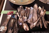 Photo showing a large pile of stainless steel forks and spoons, some wrapped in plastic, some loose, next to packaged plastic forks.