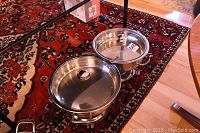 Two stainless steel cooking pots on a patterned red carpet, each with a glass lid with metal handle and metal side handles, showing the 13 inch and 12.5 inch diameter pots and their metal warming stands.