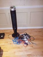 Tall black vertical PC fan with silver top on a round black base, placed on a hardwood floor against a beige paneled wall, seen with extension cords beside it.