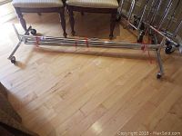 Two long chrome hanging rods bundled with red tape alongside one T-shaped base on hardwood floor
