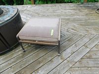 Front and top-left side view of the metal ottoman with beige cushion on a wooden deck.