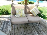 Front view of metal patio love seat with beige cushions and two patterned throw pillows on wooden deck near pool.
