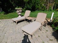 Two metal chaise loungers with beige cushions and matching small metal end table on stone patio