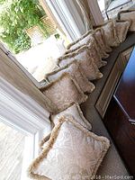Twelve beige patterned square pillows with tasseled fringe trim arranged on a cushioned window bench near large windows.