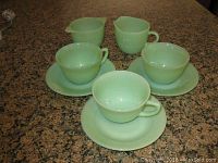 Front view of three Fire King jadeite green teacups on matching saucers and creamer and sugar bowl on granite counter