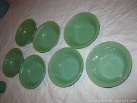 Seven green Fire King jadeite glass bowls arranged on a white quilted surface.