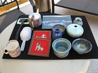 A collection of pottery items arranged on a black cloth, including a silver teapot, white pitcher, oversized coffee mug, small mirror, framed dog paintings, and multiple vases and bowls.