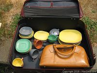 Open suitcase showing colorful Melmac dishes and brown leather handbag inside.