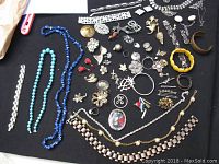 Wide view showing a large assortment of costume jewelry including multiple necklaces with turquoise and blue beads, silver-tone bracelets, rhinestone pieces, and various pins and brooches arranged on a black background.