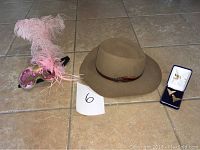 Overall lot image showing the Akubra cowboy hat, Venetian masquerade mask with feathers, and a blue presentation box with two metallic accessories.