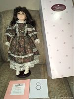 Elizabeth collectible porcelain doll standing on a dark stand wearing a brown paisley dress with lace, displayed on a sofa. Pink original box and certificate of authenticity placed in front.