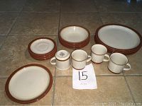 22 pieces of cream-colored stoneware dishes with brown rim, including plates, bowls, and mugs.