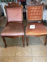 Front view of two antique Eastlake chairs side by side on tile floor, showing detailed carved wood backs and different upholstery styles.