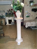 Tall white classical-style plaster pedestal with flower pot and artificial flowers on top, situated in room with tiled floor and furniture.