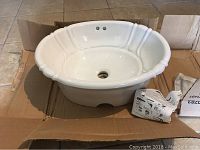 White oval ceramic drop-in sink shown inside cardboard box with instruction sheet nearby. Top and angled views capture oval shape, faucet holes, drain hole and fluted rim design.