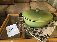 Antique green cast iron casserole with lid sitting on floral patterned cloth on a glass table, showing the full outer view including lid.