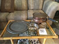 Multiple copper food warmers and a small copper colander displayed on a glass-top wicker table, alongside a large pewter and copper plate.