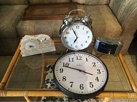 Four clocks on a glass table: marble clock, large twin bell alarm clock, Sanyo digital alarm clock, and Bates office wall clock