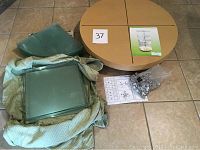 Disassembled tempered glass shelves and round wooden base and top packed in a cloth for protection, assembly hardware in a plastic bag, and assembly instructions spread out on the floor.