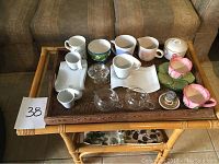 Photo of an assortment of cups, glassware, a sugar bowl, and decorative ceramic dishes on a bamboo table in front of a sofa.