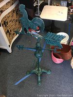Full view of the weather vane standing on the floor among other items, showing the rooster figure on top and the base.