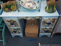 Front view of the vintage painted desk showing five drawers with seashell and coral fabric panels and blue round knobs, with a woven basket underneath.