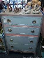 Front view of the chest showing four light blue painted drawers with pink trim and oval metal handles. Figurines and decorations rest atop the chest.