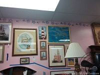 Wall showing nine pieces of coastal and lighthouse themed art hung on pink wall above lamp and furniture, featuring maritime scenes and lighthouses.