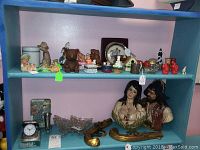 Shot of assorted figurines, trinket boxes, a clock, and decorative items arranged on two blue shelves against a pink wall.