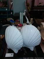 Front view of the pair of white seashell-shaped lamps showing ridged detailing, with cords visible, placed on a table surrounded by other items including a lighthouse-themed gum dispenser.