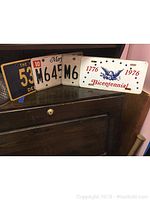 Four vintage license plates displayed on dark wood surface, showing various states and Bicentennial plate with eagle design
