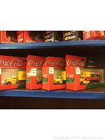 Four Coca-Cola branded die-cast trucks in original red packaging with plastic windows showing yellow trucks with red wheels and Coca-Cola branding on trailers.