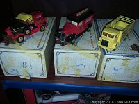Three Coca Cola collectible die-cast model trucks displayed on their original boxes. Two trucks are red with Coca Cola branding, and one is a yellow flatbed truck.