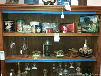 A wooden shelf holding an assortment of vintage tins, glass bottles, and three small wooden Swiss cabin themed music boxes. The tins vary in size, shape and design including a decorative bear tin and a Maine house themed tin. The glass milk bottles have wire handles. Additional vintage tins and small decor items are located around the music boxes.